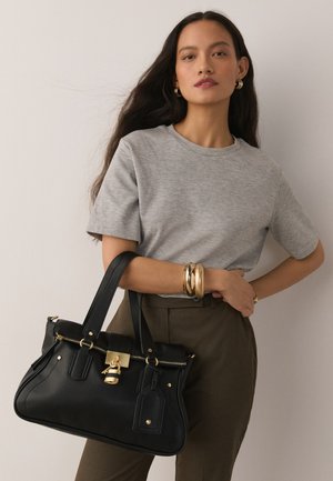 Donna che indossa una maglia grigia a maniche corte e pantaloni marroni, tiene in mano una borsa nera con chiusura dorata e indossa orecchini a cerchio dorati e braccialetti spessi.
