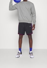 Sudadera gris, pantalones cortos negros con acentos morados, calcetines azules y zapatillas blancas con cordones multicolores. Sosteniendo un balón de baloncesto azul.