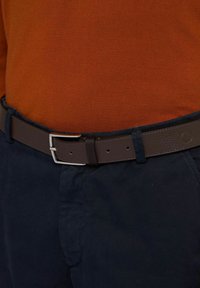 Ceinture en cuir marron avec une boucle ton argentée, portée sur un pantalon bleu marine, accompagnée d'un haut en maille orange. Design simple, accents minimalistes.