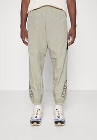 Světlé zelené, lehké sportovní kalhoty s elastickým pasem a zúženými nohavicemi. Vyznačují se černým akcentem a grafikou "JANZARUZ" na boku.