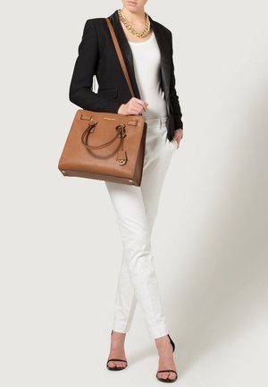 Vrouw met zwarte blazer, wit topje en broek, zwarte hakken, gouden kettingketting, draagt een bruine leren handtas over de schouder.