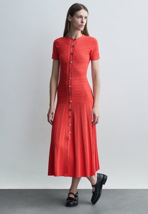 Mujer con un vestido midi rojo texturizado de botones y mangas cortas, y mocasines negros, posando frente a un fondo claro y liso.