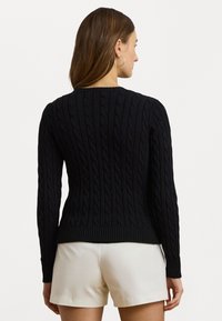 Pull noir en maille torsadée avec des manches longues, des poignets et un bord-côte, agrémenté d'un motif texturé. Porté avec un short de couleur claire.
