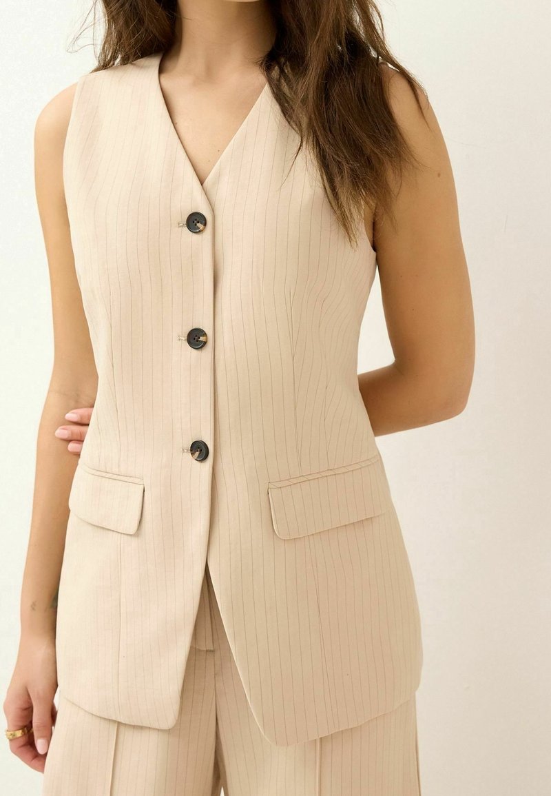 Femme portant un gilet sans manches beige à fines rayures avec trois boutons noirs et un pantalon assorti sur un fond uni.