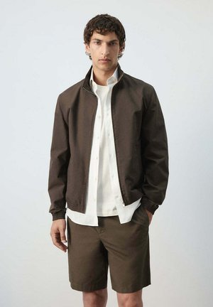 Junger Mann steht und trägt eine dunkelbraune Jacke mit Reißverschluss, ein weißes Hemd mit Knöpfen, ein weißes T-Shirt und passende dunkelbraune Shorts vor einem einfarbigen Hintergrund.