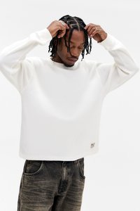 Fehér bordázott pulóver kerek nyakkivágással és hosszú ujjakkal, az alsó szegély közelében egy kis logófolttal. Sötét farmernadrággal párosítva.