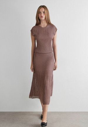 Frau mit langen, glatten Haaren, die ein ärmelloses Stricktop und einen passenden Midirock in staubigem Rosa trägt, steht vor einer schlichten weißen Wand.