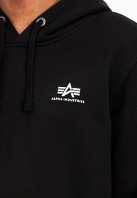 Felpa nera con cappuccio regolabile, polsini a coste e logo bianco ricamato sul lato sinistro del petto. Tessuto morbido.