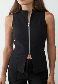 Zwarte mouwloze top met een hoge kraag en voorkleurige rits, met een strakke pasvorm en split aan de onderzijde. Glad textuur, minimalistische stijl.