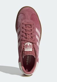 Růžové semišové tenisky s bílými pruhy a texturovaným jazykem. Mají gumovou podrážku, zaoblenou špičku a detail loga na jazyku.