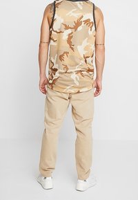 Débardeur en maille beige avec un motif camouflage marron, associé à un pantalon beige. Les chaussures sont des baskets blanches. Design minimaliste et coupe décontractée.