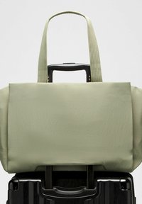 Zöld bőr bevásárlótáska textúrált felülettel, hosszú fülekkel és  bőrönd fogantyújának, valamint egy fekete kerekes bőröndön elhelyezett részel.