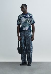 Μπλε πουλόβερ με σχέδια και πράσινες λεπτομέρειες, σε συνδυασμό με φαρδιά, υφασμάτινη μπλε παντελόνα με μοτίβο καρφίτσας. Μαύρη τσάντα και παπούτσια ολοκληρώνουν την εμφάνιση.