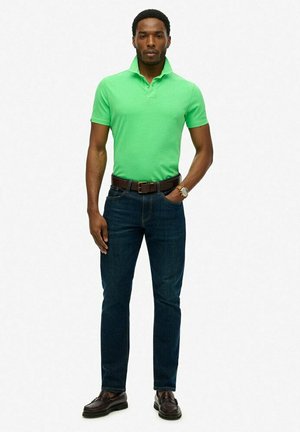 Grünes Polohemd mit kurzen Ärmeln, kombiniert mit dunkelblauen Jeans. Ein brauer Gürtel und Schuhe vervollständigen das Outfit. Einfaches Design, glatte Textur.