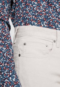 Koszula z długim rękawem w kwiatowy wzór w kolorach niebieskim, czerwonym i białym; zestawiona z jasnobeżowymi spodniami sztruksowymi o subtelnej fakturze i kieszeniach.