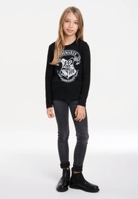 Schwarzes Langarmshirt mit dem Hogwarts-Wappen, kombiniert mit dunkelgrauen Jeans und schwarzen Stiefeletten. Schlichtes Design mit glatter Textur.