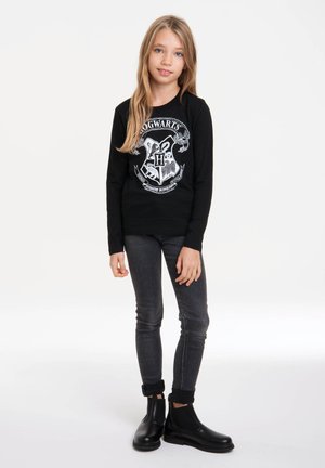 Chemise noire à manches longues avec un blason de Poudlard, assortie à un jean gris foncé et des bottines noires. Design simple, avec une texture lisse.