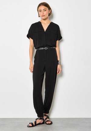 Donna in piedi contro un muro bianco che indossa una tuta nera a maniche corte, cintura nera, braccialetto a polsino argentato e sandali neri.