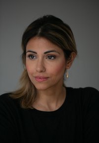 Porträt einer Person mit langen, dunklen Haaren und einem schwarzen Oberteil. Sie trägt einen goldenen Tropfenohrring und hat dezentes Make-up mit einem natürlichen Look.
