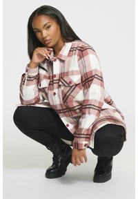 Koszula flanelowa w różowo-białą kratę z zapięciem na guziki, kieszeniami na piersi i kontrastującymi guzikami, połączona z czarnymi legginsami i butami. 