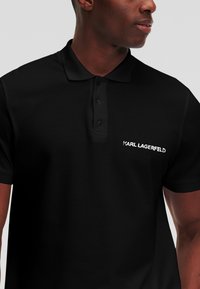 Črna polo majica iz mehkega materiala, s tri gumbi na zapenjanju in izvezenim logotipom "KARL LAGERFELD" na levem prsnem delu.