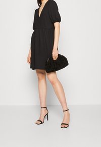 Vestido negro de manga abullonada con escote en V profundo, cintura ajustada, combinado con tacones negros con tiras y un bolso negro texturizado con asa.