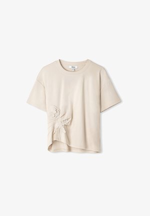 Béžové tričko s krátkým rukávem a zakřiveným lemem, se strukturovanou květinovou výšivkou na levé straně. Vyrobeno z měkké bavlněné tkaniny.