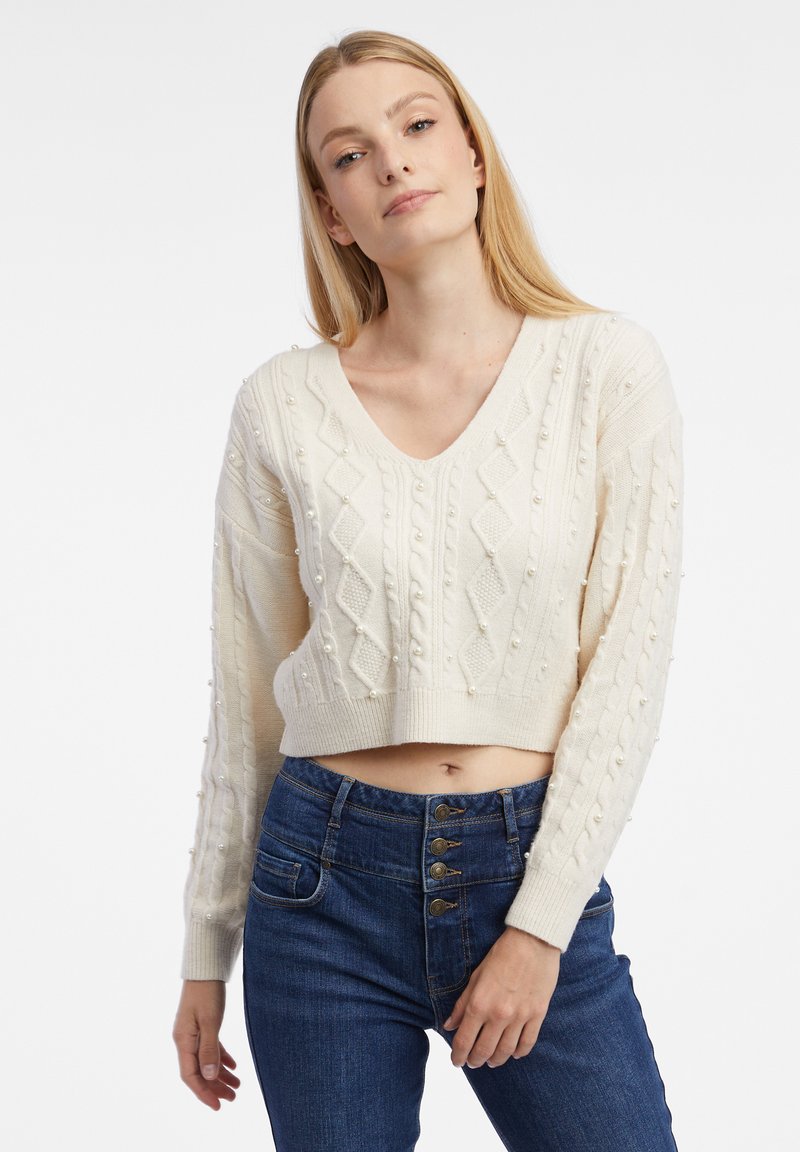 Pull en tricot blanc avec un col en V, présentant des motifs en losange texturés et des accents de perles surélevées, associé à un jean bleu taille haute.