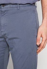 Gros plan sur une personne portant un pantalon bleu avec un bouton et une poche latérale, et une chemise bleue rentrée, la main reposant le long du corps.