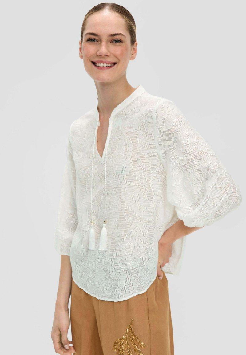 Lächelnde Frau in einer durchsichtigen weißen Bluse mit Muster und Quastenbändern, kombiniert mit braunen Hosen mit goldenen Blattformen, vor einem schlichten Hintergrund stehend.