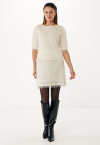 Jasnobeżowa sukienka swetrowa z frędzlami, z rękawami 3/4, fakturowaną powierzchnią i frędzlami na hemie. Noszona z czarnymi, sięgającymi do kolan botkami.