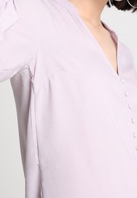 Blusa lilla chiaro con scollo a V, dettagli con bottoni lungo il davanti, maniche a sbuffo e tessuto texturizzato.