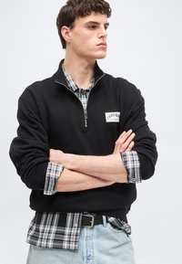 Νεαρός άνδρας με σταυρωμένα χέρια, φοράει μαύρο πουλόβερ Calvin Klein με φερμουάρ στο λαιμό πάνω από μια ασπρόμαυρη καρό πουκάμισο και ανοιχτόχρωμα γαλάζια τζιν.