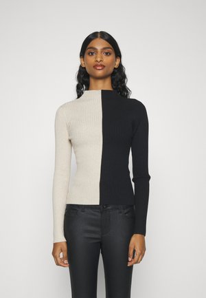 Ribbet langermet topp med høy krage, med delt design: den ene siden beige, den andre siden svart, kombinert med svarte bukser.