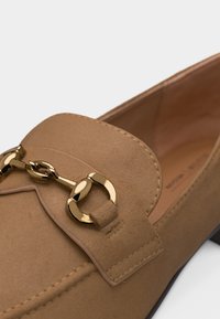 Mocassin en daim tan avec un détail en métal doré en forme de mors de cheval sur le dessus, présentant une couture fine et un petit talon noir.