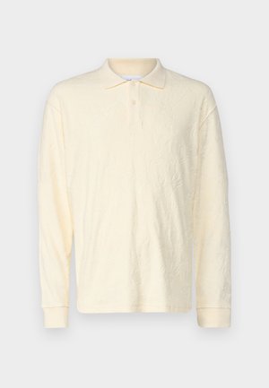 Polo majica s dugim rukavima od mekog, teksturiranog kremastog materijala. Ima klasičan ovratnik, dva dugmeta i rebraste manžete.