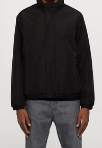 Černá bunda na zip s vysokým límcem, elastickými manžetami a lemem. Vyrobená z hladké, voděodolné látky. Přední kapsy jsou viditelné.