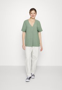 Grønn V-hals topp med korte ermer, lett tekstur, kombinert med hvite bukser og svart-hvite sneakers, mot en ensfarget bakgrunn.
