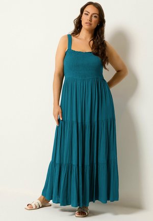 Frau mit langen dunklen Haaren, die ein ärmelloses, türkisfarbenes gestuftes Maxikleid und weiße Slide-Sandalen trägt, steht vor einem schlichten hellen Hintergrund.