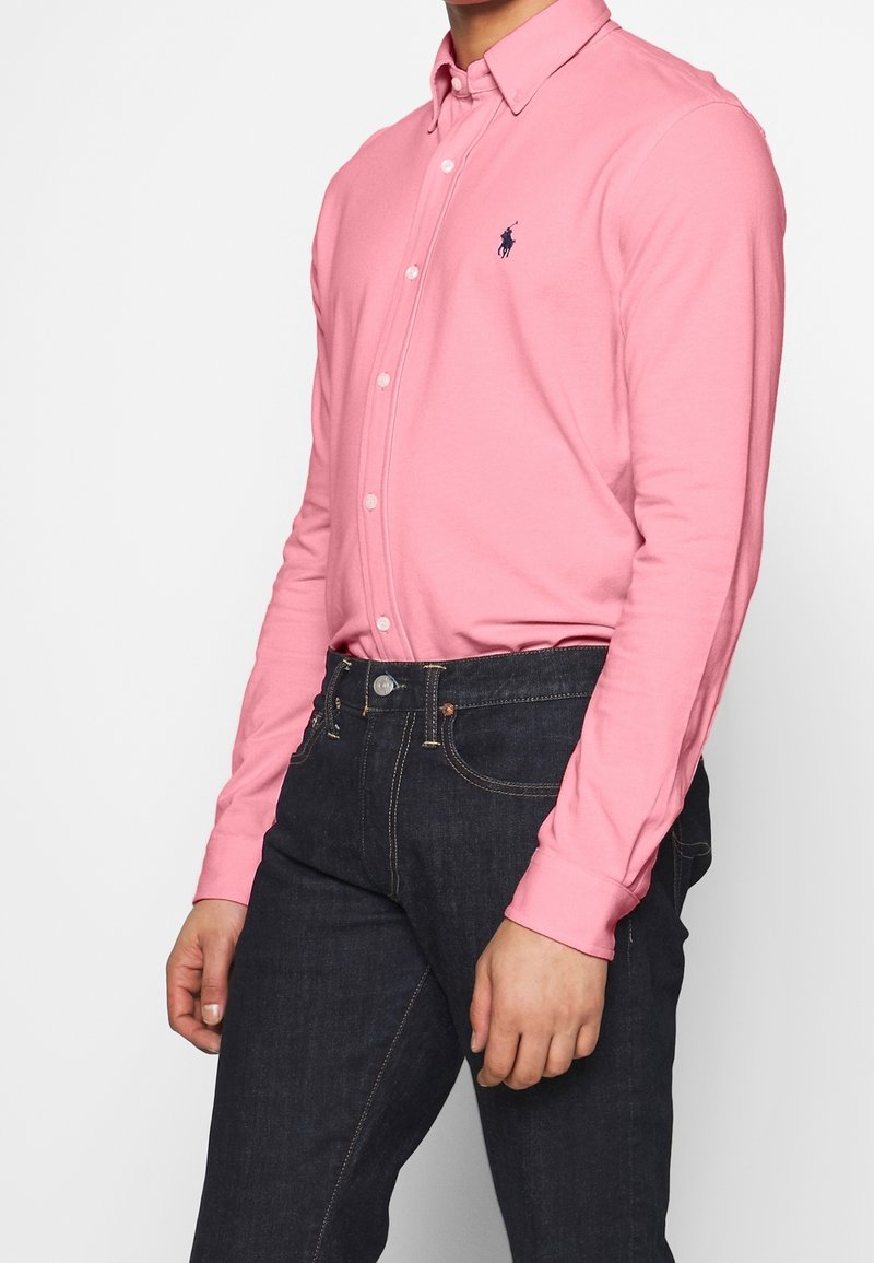 Muž v růžové košili s dlouhým rukávem a modrým logem, v tmavě modrých džínách, zobrazen od hrudníku po střed stehen.