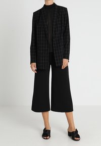 Black and white checked blazer over a sheer black top, paired with wide black culottes and black slide sandals with bow accents.