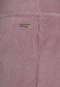 Tissu en velours côtelé rose avec une texture côtelée, arborant une petite étiquette logo dorée. La surface est lisse et douce, avec des détails de couture visibles.