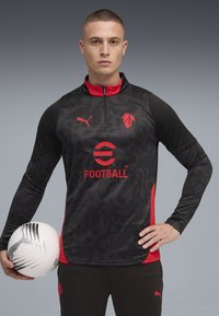 Haut d'entraînement de football à manches longues noir et rouge avec un col zippé, présentant un motif texturé et un logo sur la poitrine, accompagné d'un ballon de football blanc.