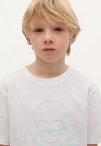 T-shirt bianco in cotone con un design di teschio e croce di ossa azzurro chiaro sul davanti. La maglietta ha un colletto rotondo e maniche corte.