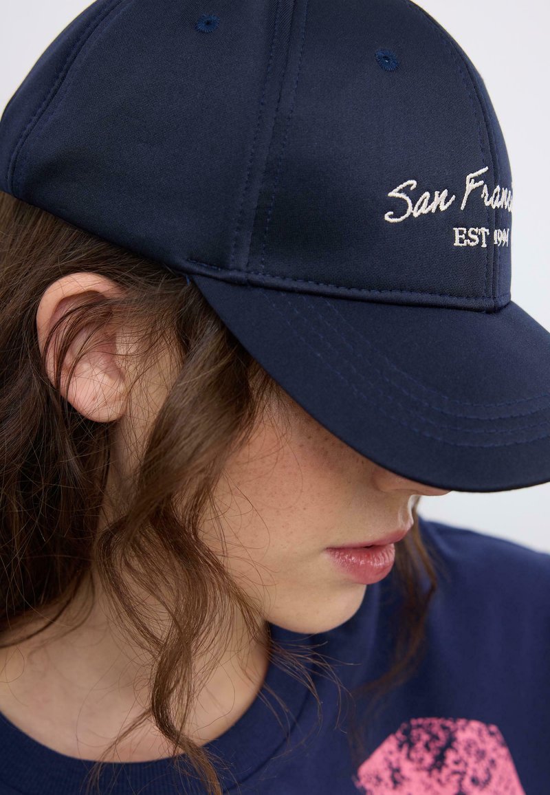 Giovane con capelli castani ondulati indossa un berretto blu navy ricamato con la scritta "San Francisco EST 1991" e una maglietta blu navy con un motivo rosa.