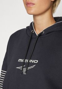 Marinehue med hvite snorer og logo. Har stripete mønster på ermene, laget av en blanding av bomull for en myk tekstur.