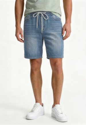 Muž nosí svetlomodré denimové šortky s bielou šnúrkou a bielymi teniskami, stojí na jednoliatom pozadí.