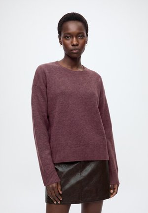 Femme aux cheveux courts portant un pull violet et une jupe en cuir noire, debout devant un fond clair uni.