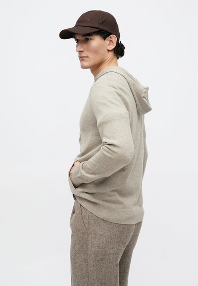 Beige hoodie med snoretræk i hætten, strikket tekstur og kengurulomme, parret med brune strukturerede bukser og en brun baseballkasket.
