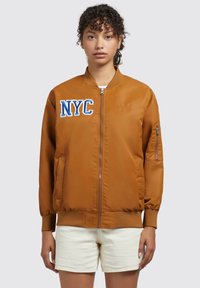 Giubbotto bomber marrone con polsini a costine, chiusura frontale con zip, ricamo NYC in blu e bianco sul petto, e tasche laterali. Tessuto di superficie morbida.